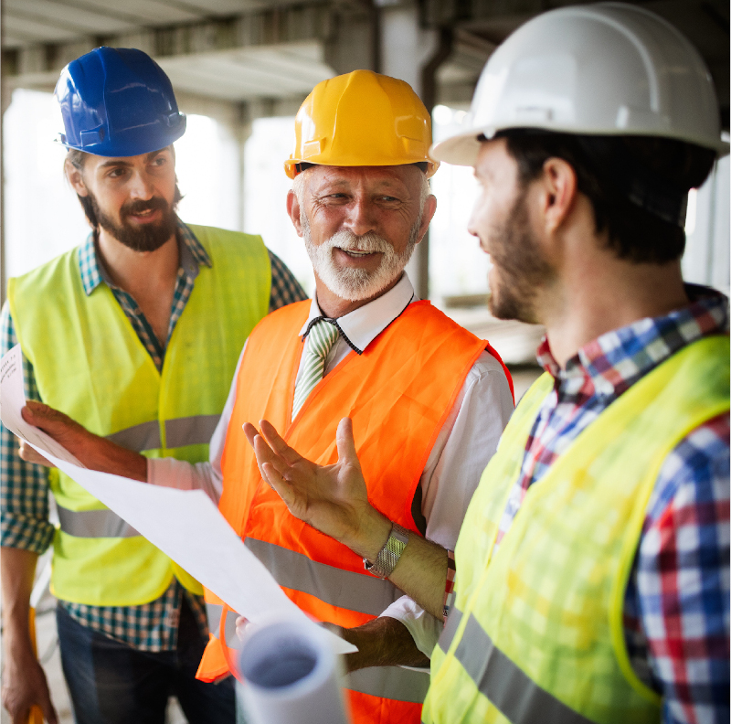Three construction workers onsite in safety gear discussing blueprints