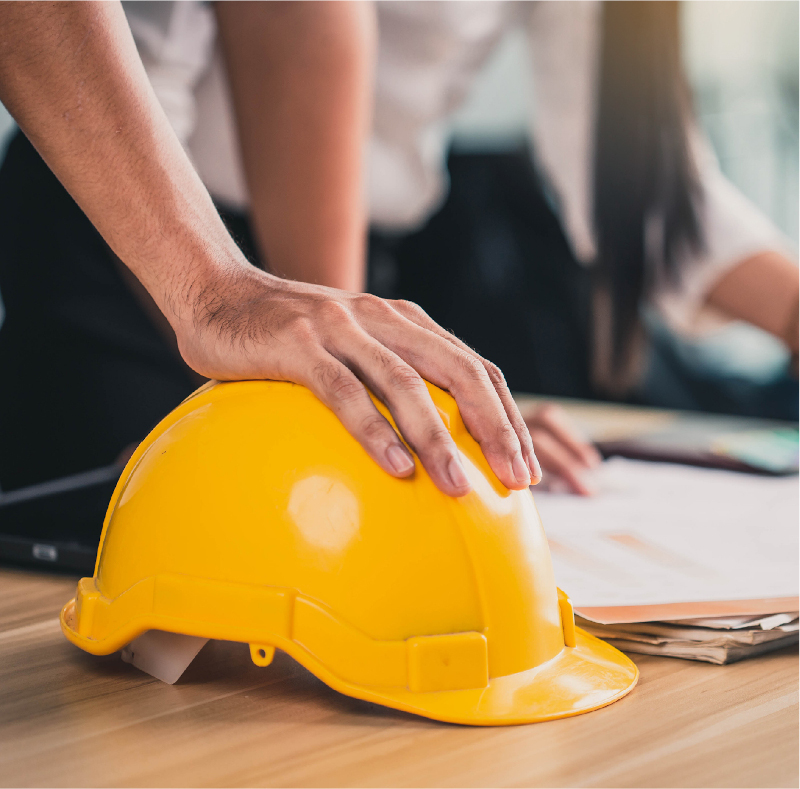 Man's hand resting on a yellow hard hat on a drafting desk